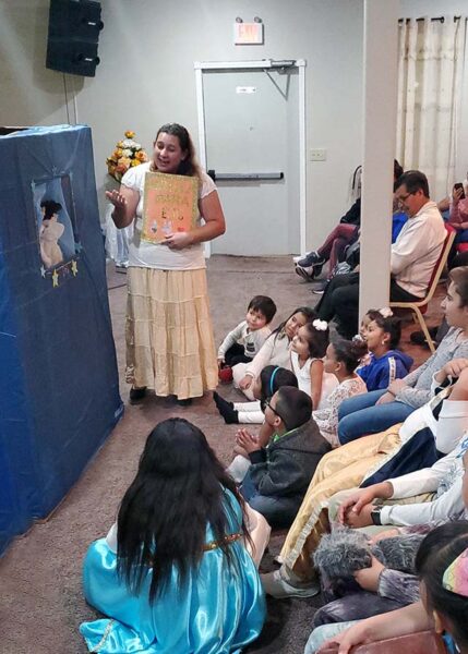 Children gathered around a puppet show near the stage while a woman narrates a Bible story.